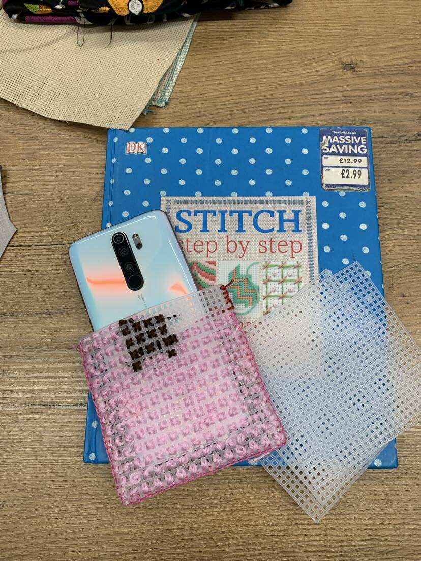 Na drewnianym blacie leży książka „Stitch Step by Step” w niebieskiej okładce w białe kropki. Na niej znajduje się smartfon w jasnej obudowie częściowo wsunięty w różowy, plastikowy, kanwasowy pokrowiec do haftu krzyżykowego z zaczętą różowo-brązową robótką. Obok leżą dwie plastikowe siatki kanwy do haftu — jedna różowa, druga przezroczysta.