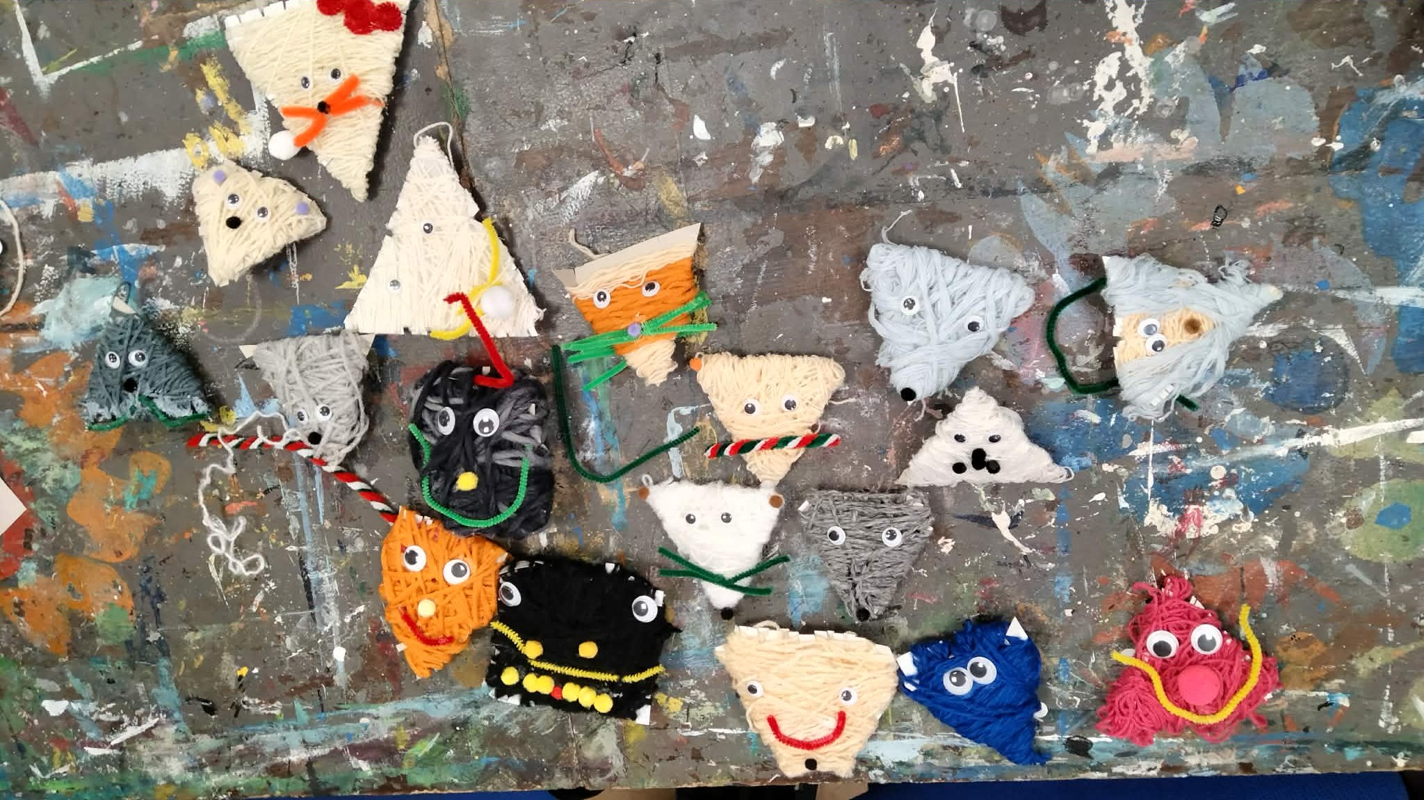 Na stole leżą prace plastyczne wykonane przez dzieci z włóczki, to kolorowe - trójkątne i kwadratowe - buźki, które mają oczy i wąsy.