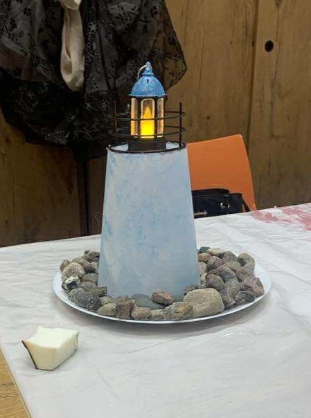 Lampion w kształcie latarni morskiej - ma białą podstawę i niebieski dach. Latarnia stoi na talerzyku wypełnionym kamieniami. Całość ustawiona na białym stole, w tle widać pomarańczowe krzesło i szafę.