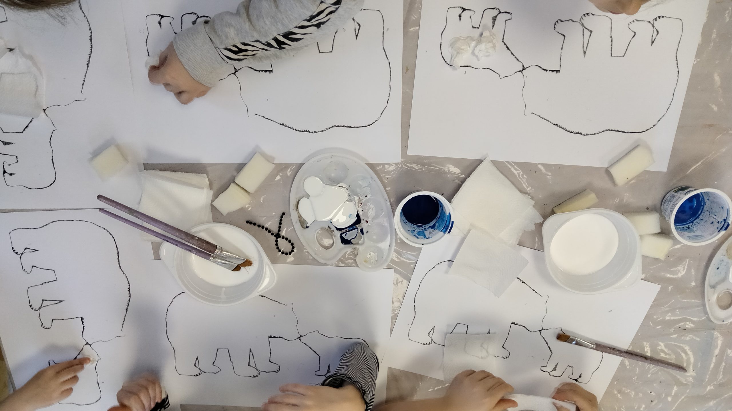 Na zdjęciu widać białe kartki z nadrukowanymi sylwetkami niedźwiedzi polarnych, dziecięce ręce oraz pojemniki z farbą, klejem i pędzle.