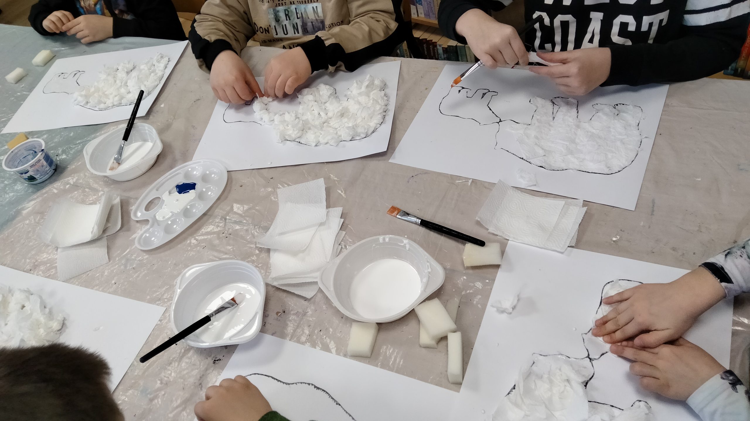 Na stole leża białe kartki z nadrukowanymi sylwetkami niedźwiedzi, dziecięce ręce oraz miseczki z klejem i pędzle.