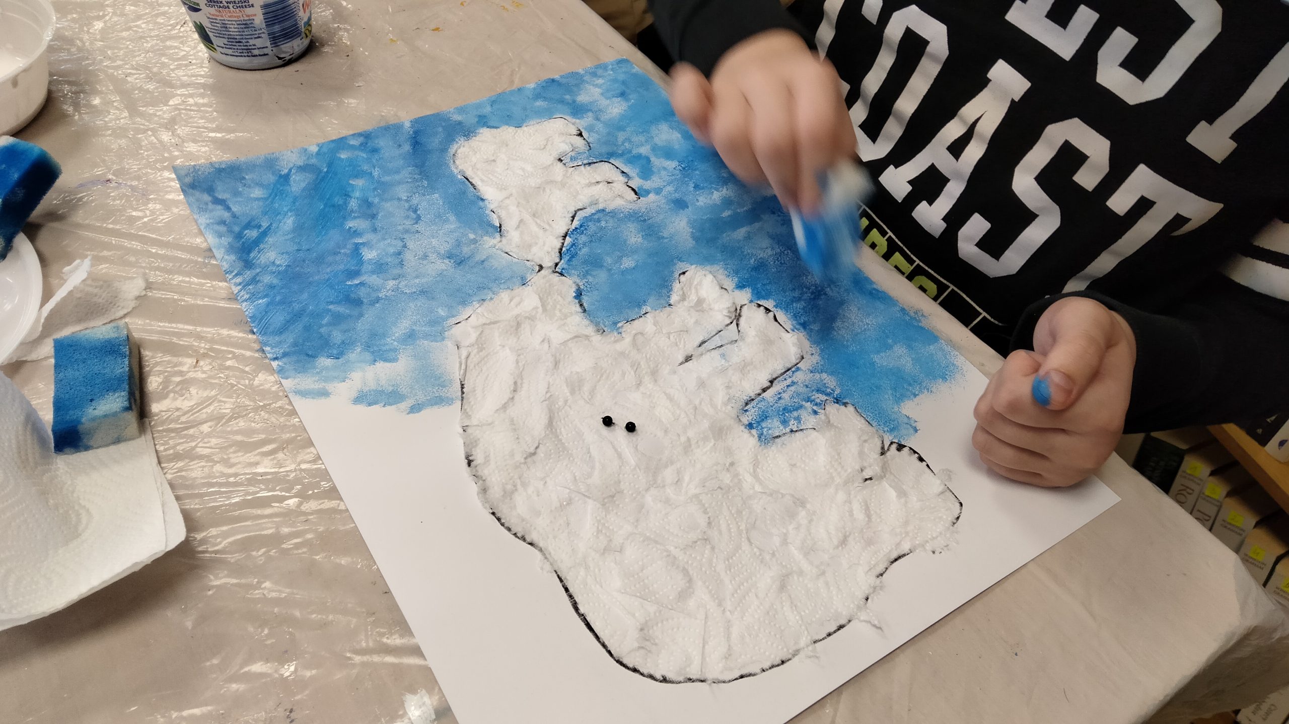 Zdjęcie przedstawia pracę plastyczną, która jest w trakcie wykonywania. Przedstawia ona dwa niedźwiedzie polarne - dużego i małego. Widać też dziecięce dłonie wykonujące niebieskie tło.