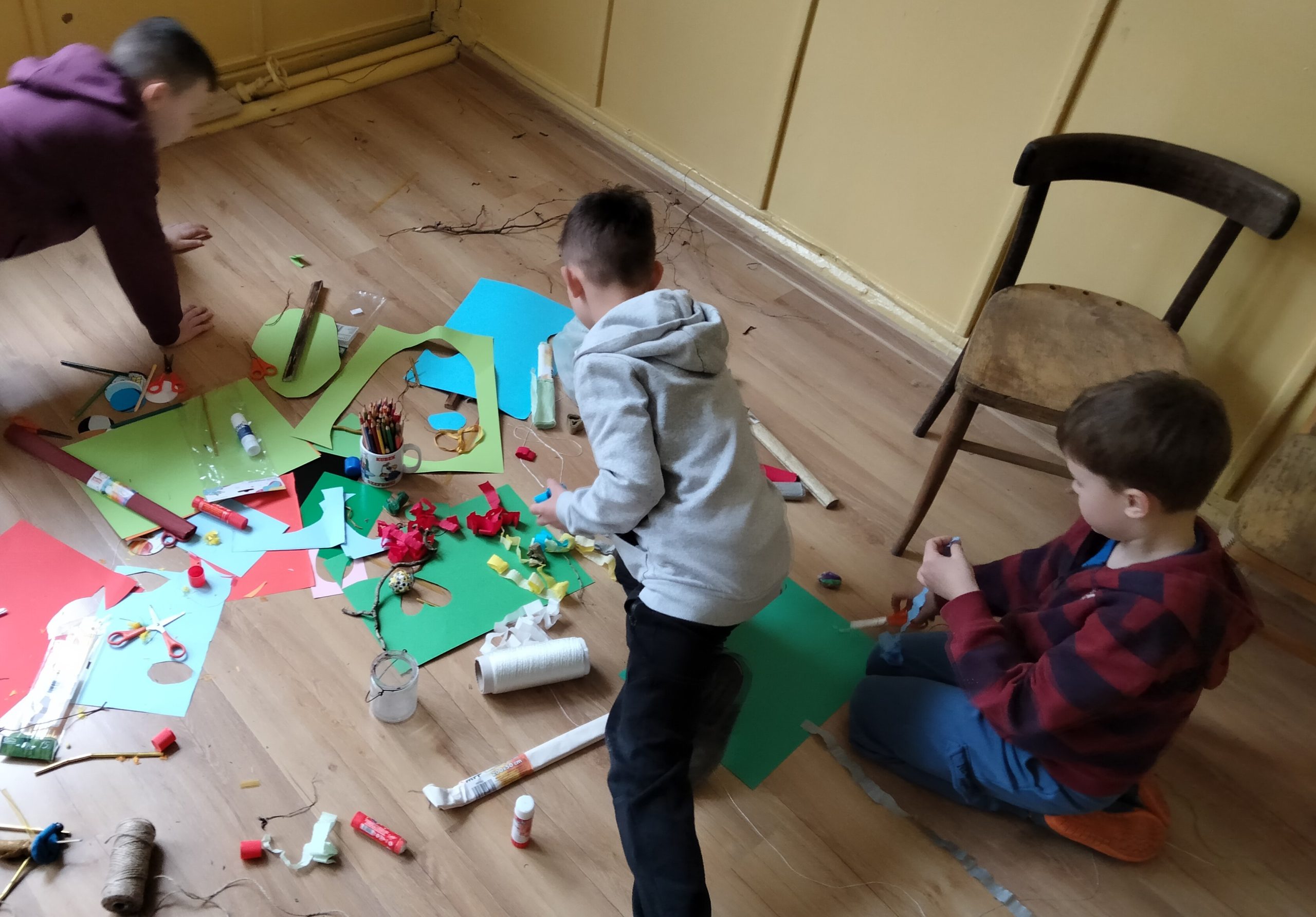 Chłopiec pochyla się nad podłogą, na której leży papier kolorowy i inne przedmioty potrzebne do prac plastycznych. Na podłodze siedzi dwóch innych chłopców.