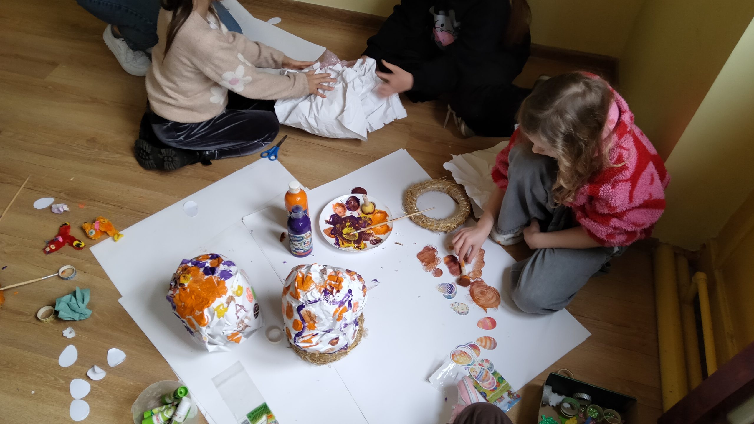 Dwoje dzieci na podłodze zgniata wielką kulę papieru, jedno z dzieci siedzi obok przed elementami pomalowanym farbami i materiałami plastycznymi.
