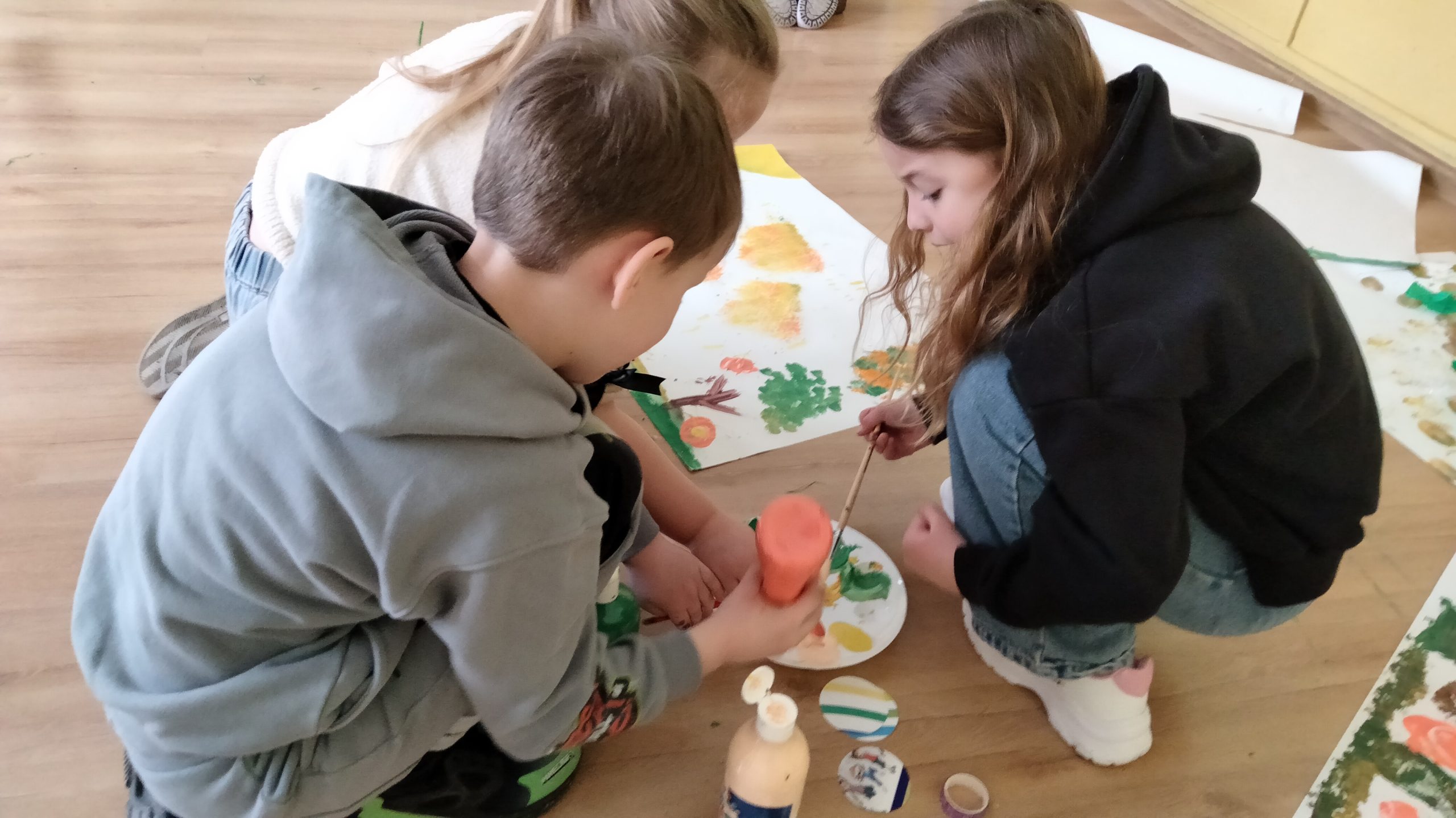 Troje dzieci zajętych farbami - jedno z dzieci wyciska farbę na paletkę, jedno trzyma pędzel.