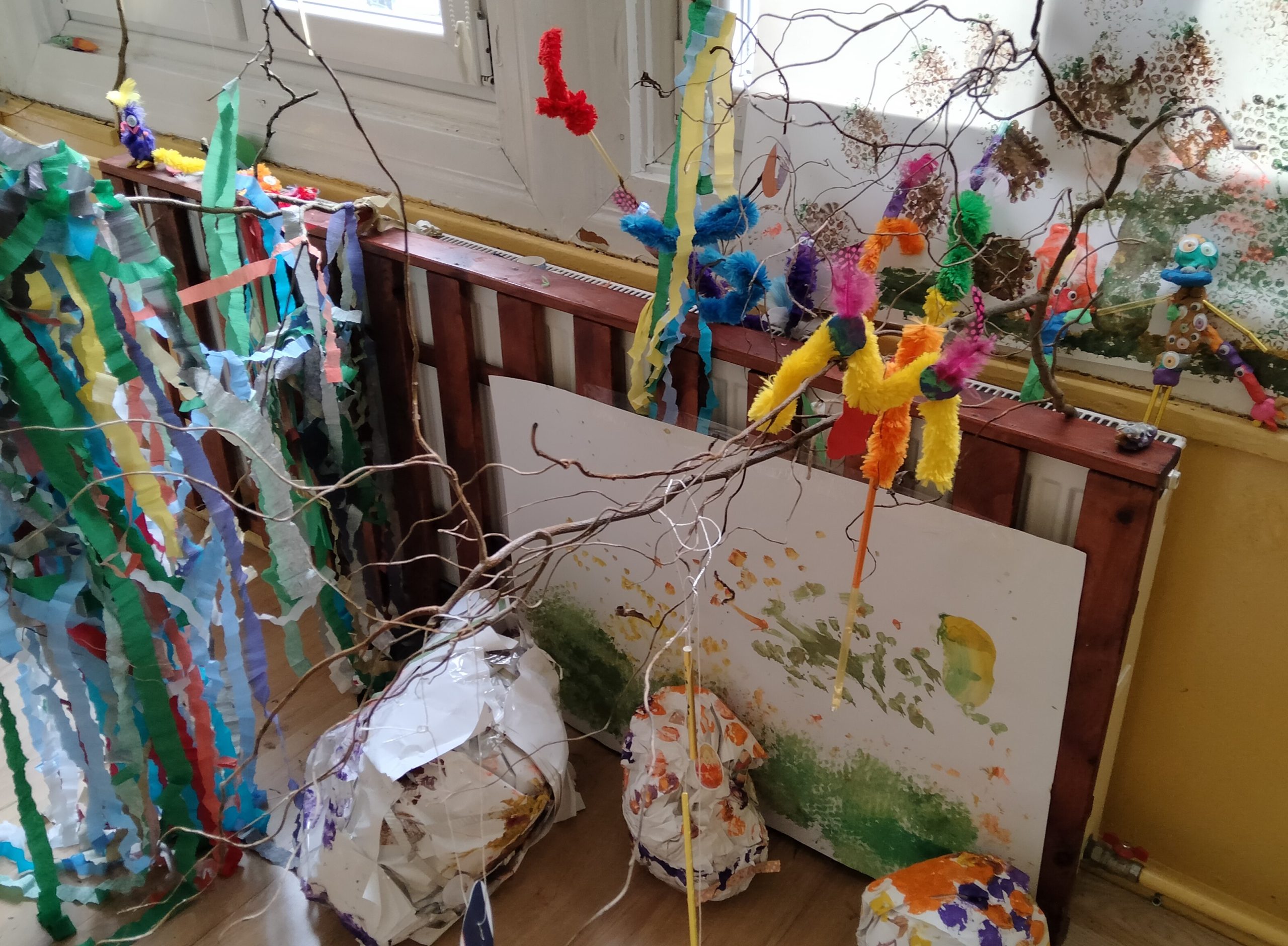 Instalacja artystyczna składająca się z gałęzi, kolorowej bibuły i dużych kartek pomalowanych farbami.