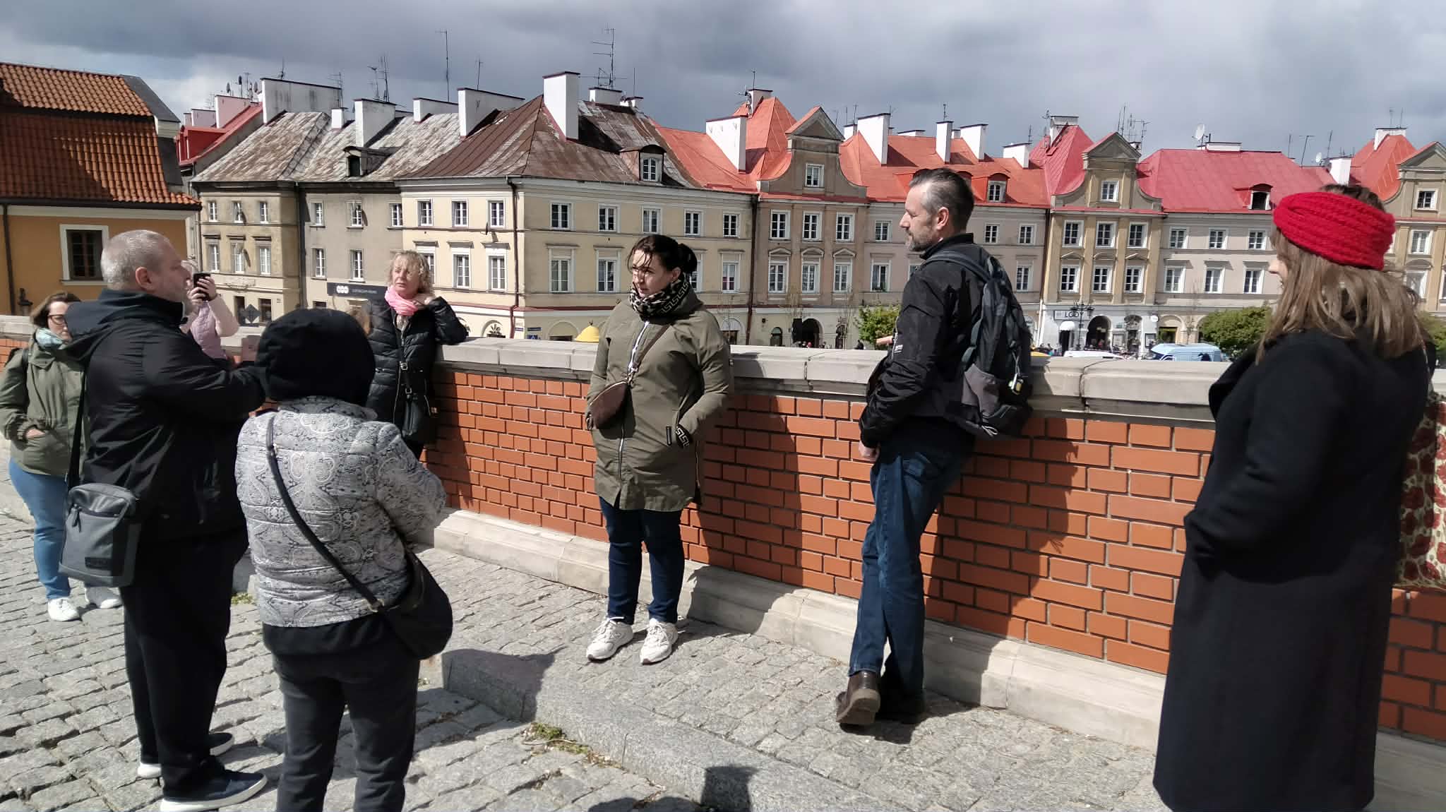 Grupa osób stoi przy ceglanym murku, w tle widać stare kamienice.