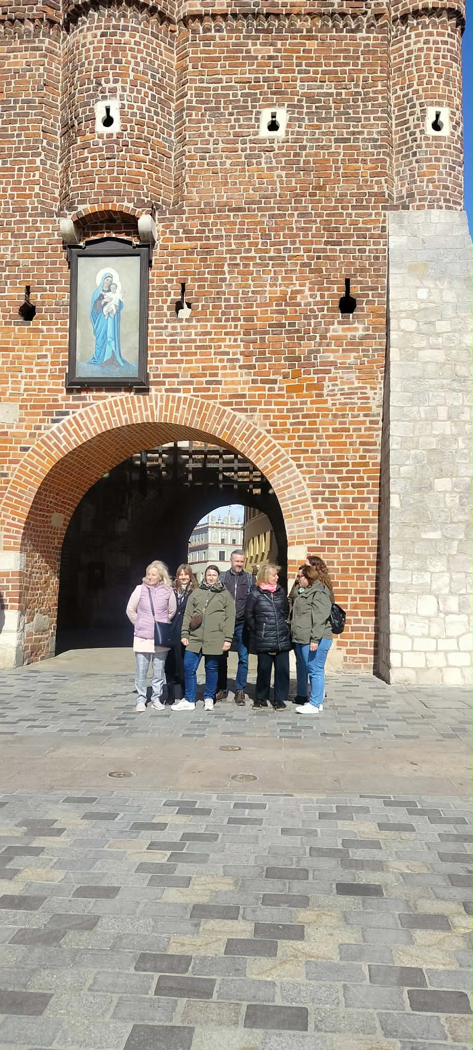Grupa osób stojąca na tle dużego wejście/bramy, która zbudowana jest z cegieł.