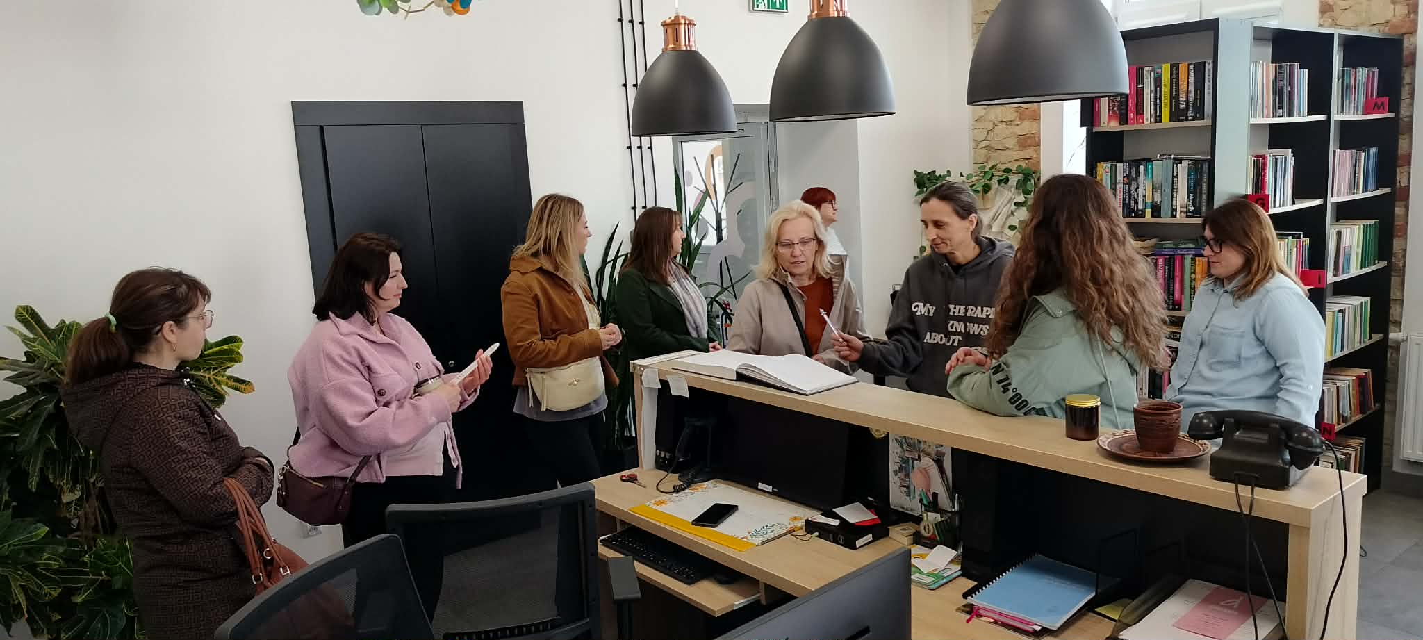 Grupa osób stojących obok otwartej książki, u góry wiszą lampy, w dolnej części zdjęcia widać biurko i krzesło.
