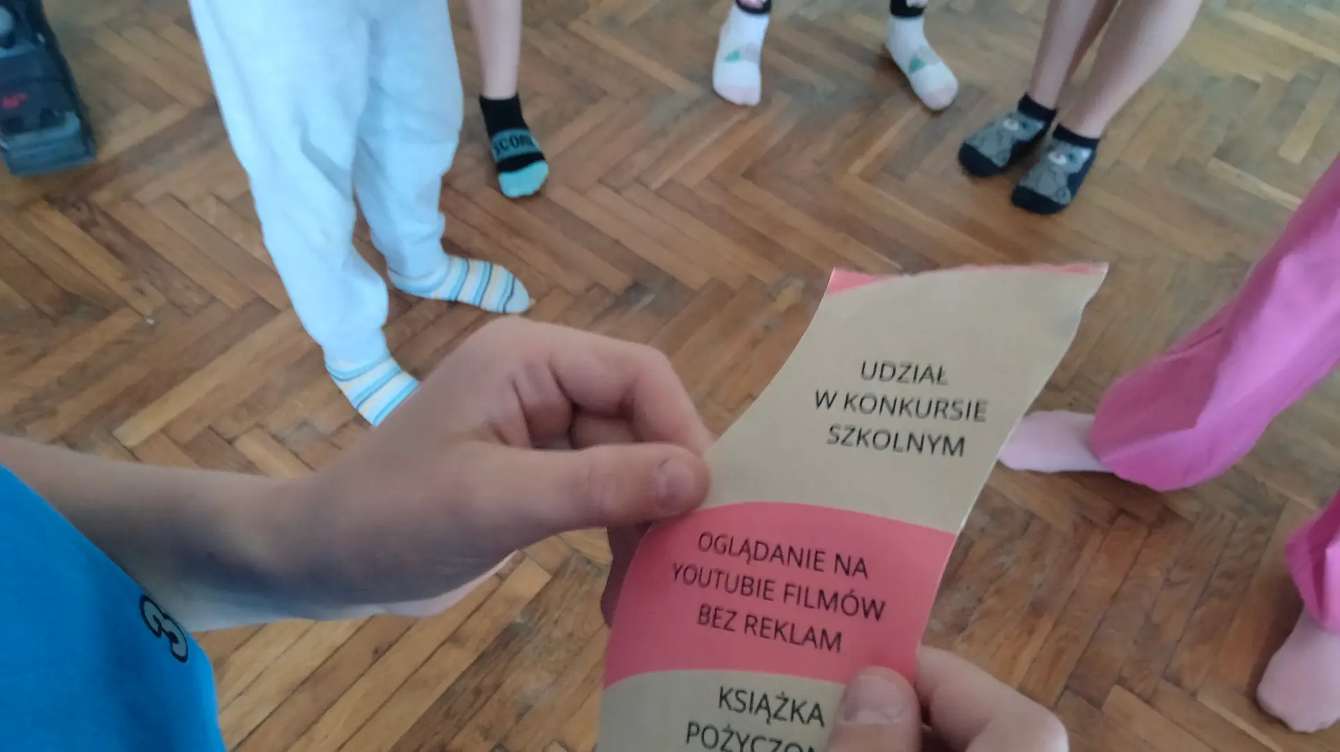 Na pierwszym planie widać fragment kartki w dłoniach. Na niej napisane są hasła na różnych kolorach, te hasła to: 