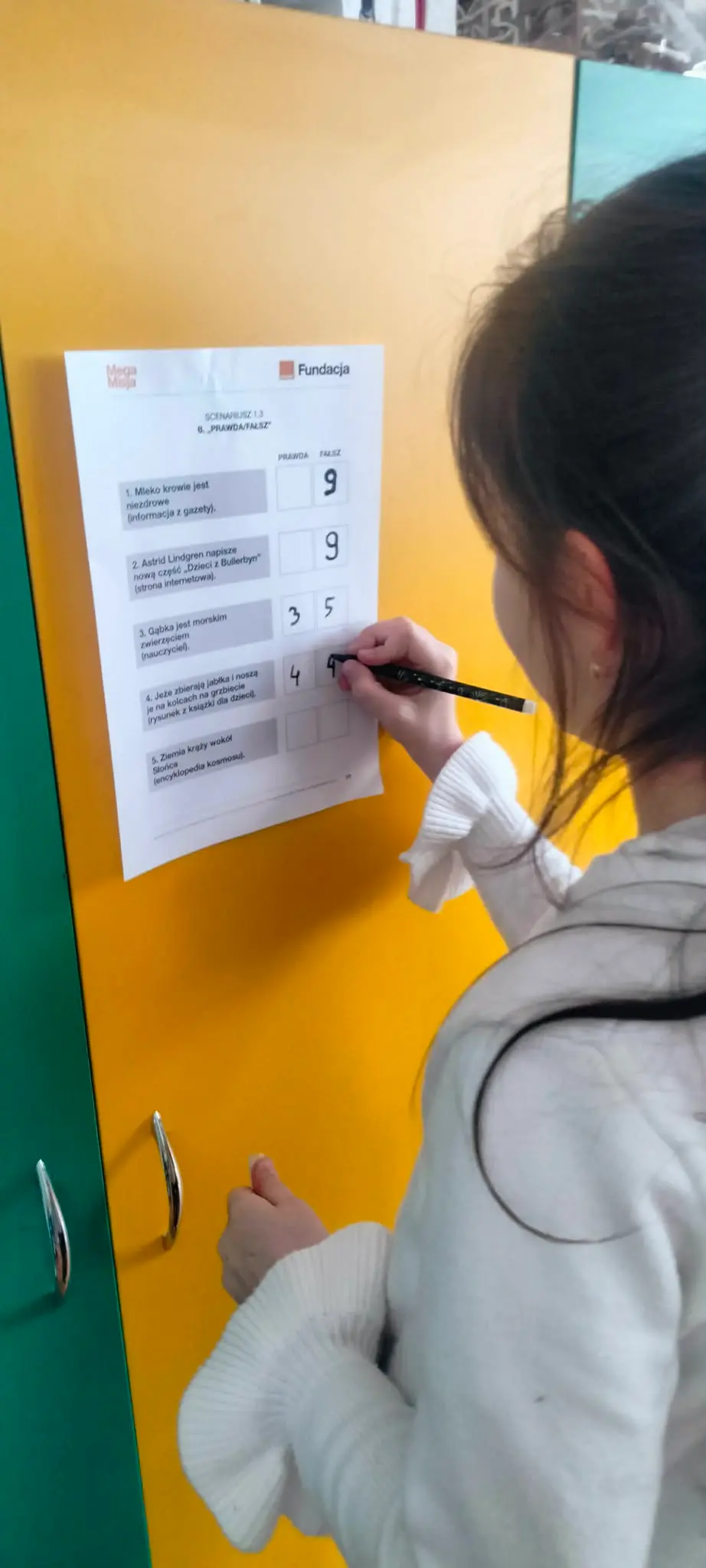 Zdjęcie przedstawia dziewczynkę, która zaznacza coś na kartce przyklejonej do drzwi.