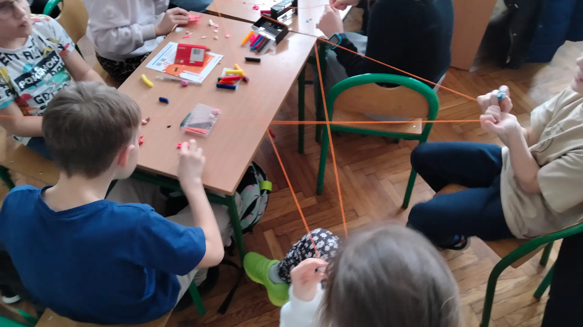 Przy stoliku dookoła siedzą dzieci. Na stoliku widać plastelinę i kartki. Osoby siedzące wokół trzymają rozciągniętą między sobą pomarańczową włóczkę.