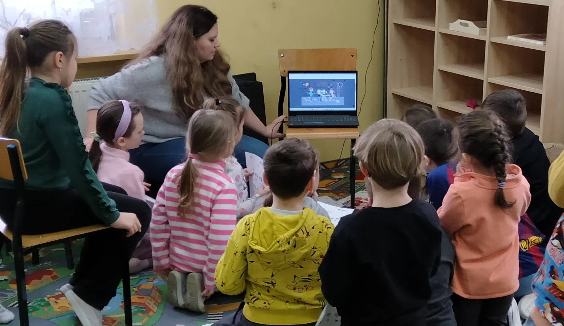 Po lewej stronie widać kobietę, która pokazuje grupie dzieci obrazek w komputerze.