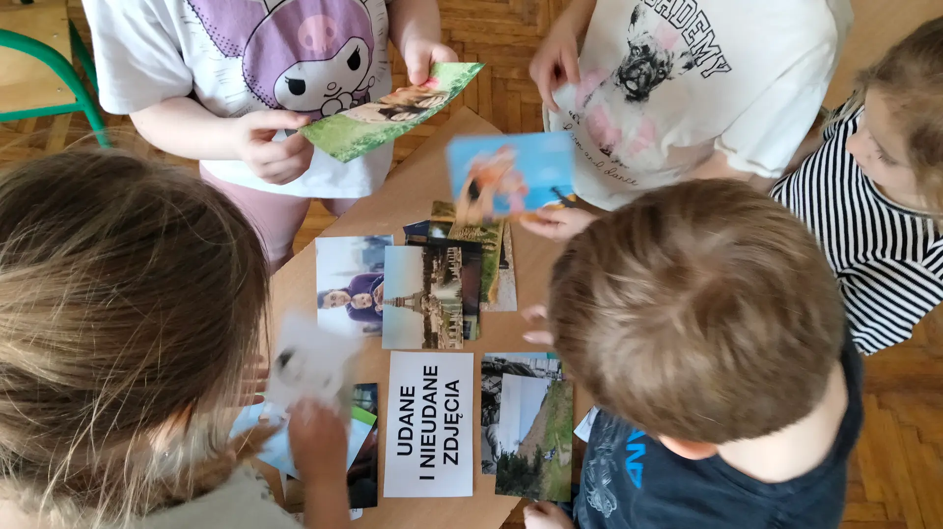 Zdjęcie pokazuje z góry grupę dzieci siedzących wokół stolika. Na stoliku leżą fotografie oraz kartka z napisem 