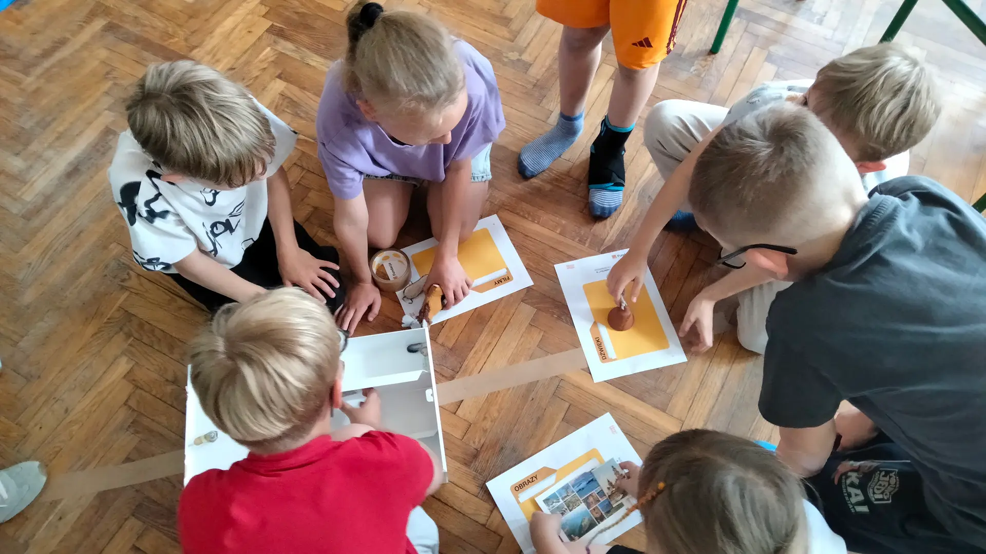 Zdjęcie przedstawia widzianą z góry grupę dzieci siedzących na podłodze. Dzieci mają przed sobą cztery kolorowe kartki i pocztówkę.