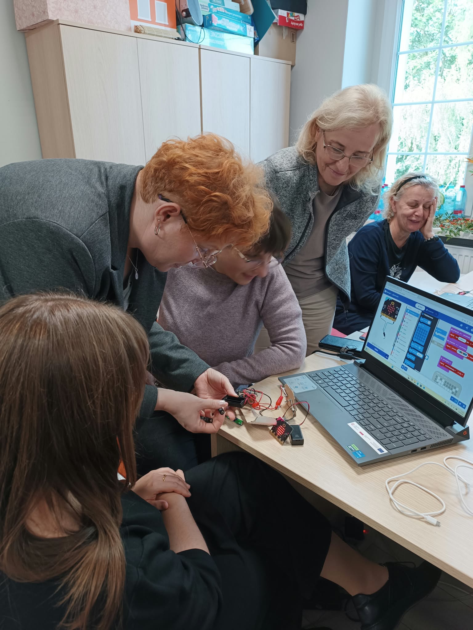 W centralnej części znajduje się kobieta, która pochyla się nad laptopem i trzyma w rękach elektroniczny element. Wokół niej znajdują się osoby, które patrzą na to, co robi.
