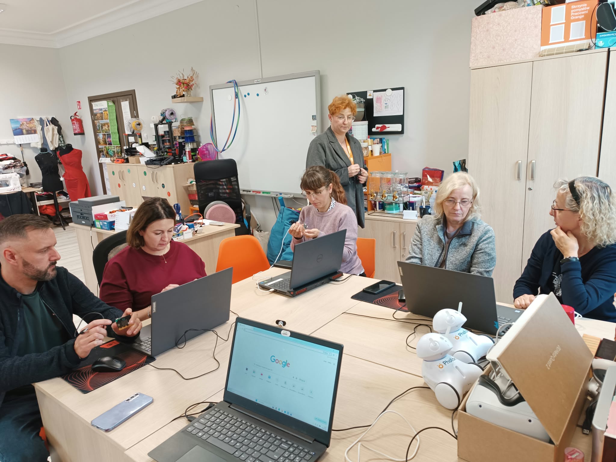 Na zdjęciu znajduje się pięć osób siedzących przy laptopach. Z tyłu, za stołem stoi kobiet, która przygląda się pracy siedzących osób. W tle na ścianie widać wiszącą białą tablicę.