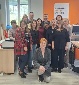 Na zdjęciu znajduje się grupa osób pozujących do zdjęcia. W tle widać okno i fragment pomarańczowej ściany z napisem "Pracownia Orange w Porębie".