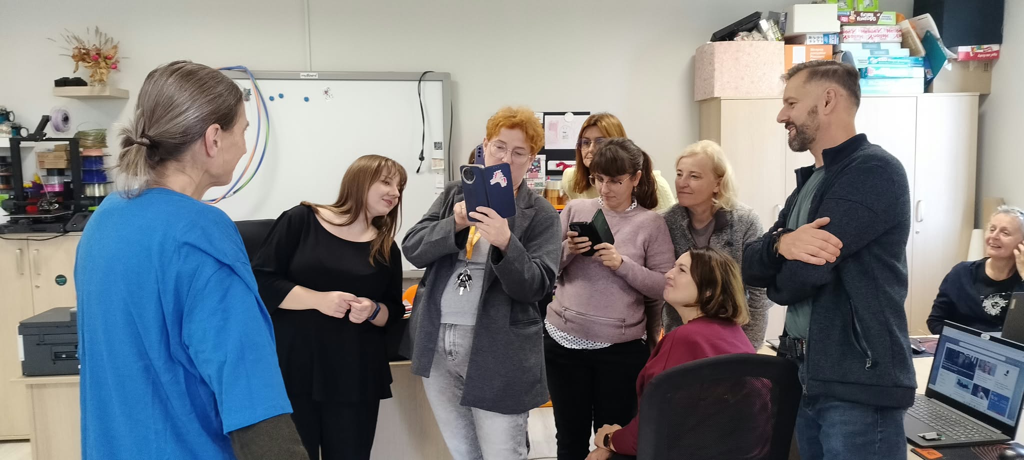 Z lewej strony stoi kobieta w niebieskiej koszulce. W prawej części zdjęcia stoi kilka osób z telefonami zwróconymi w kierunku kobiety w niebieskiej koszulce, patrzą w obiektywy i się uśmiechają.