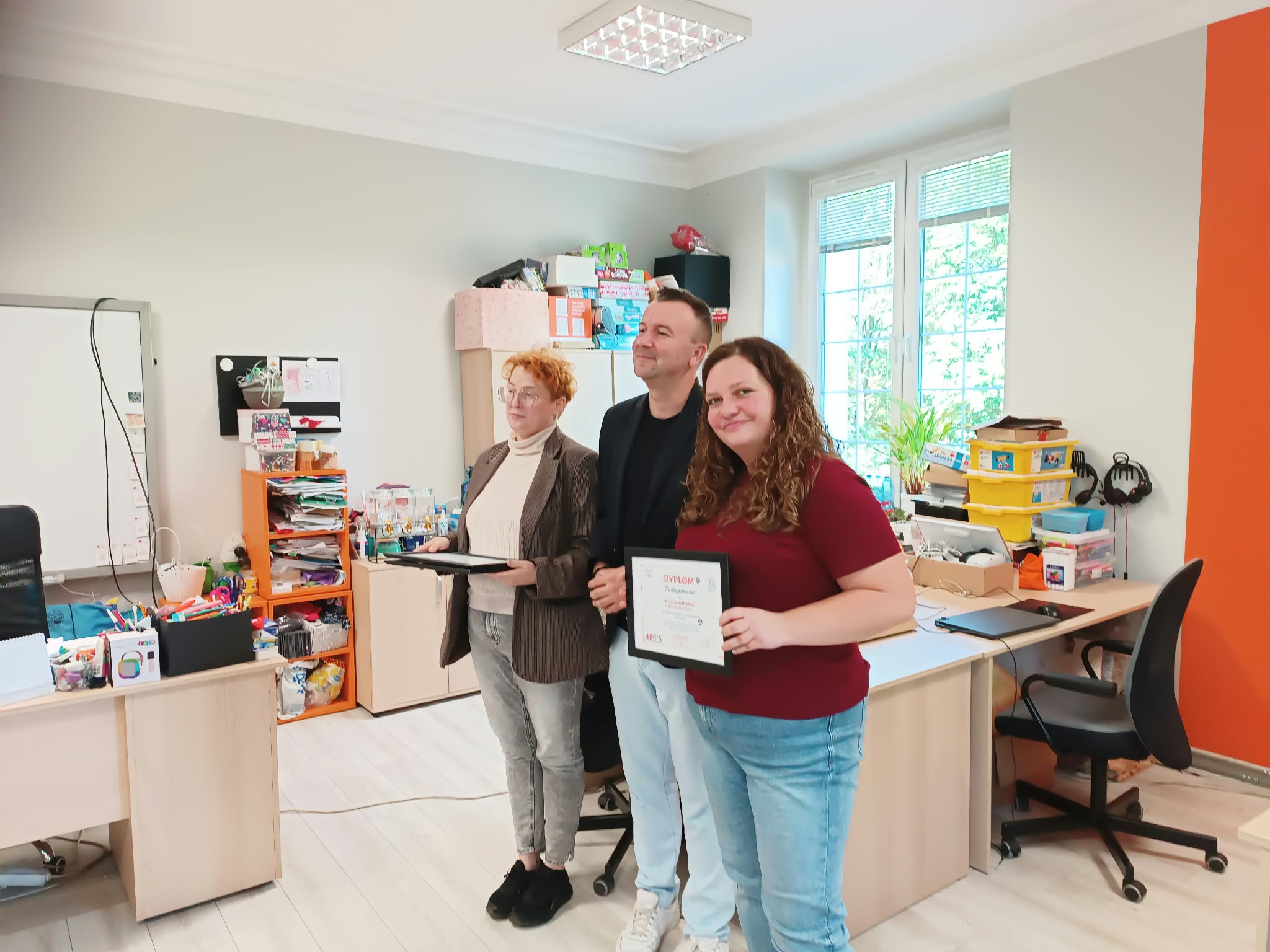 W centralnej części zdjęcia znajdują się trzy osoby, pozują do zdjęcia. Jedna z osób - kobieta - trzyma w ramce dyplom, uśmiecha się do obiektywu. W tle znajdują się półki, na których są różne przedmioty, biurko z laptopami oraz okno.