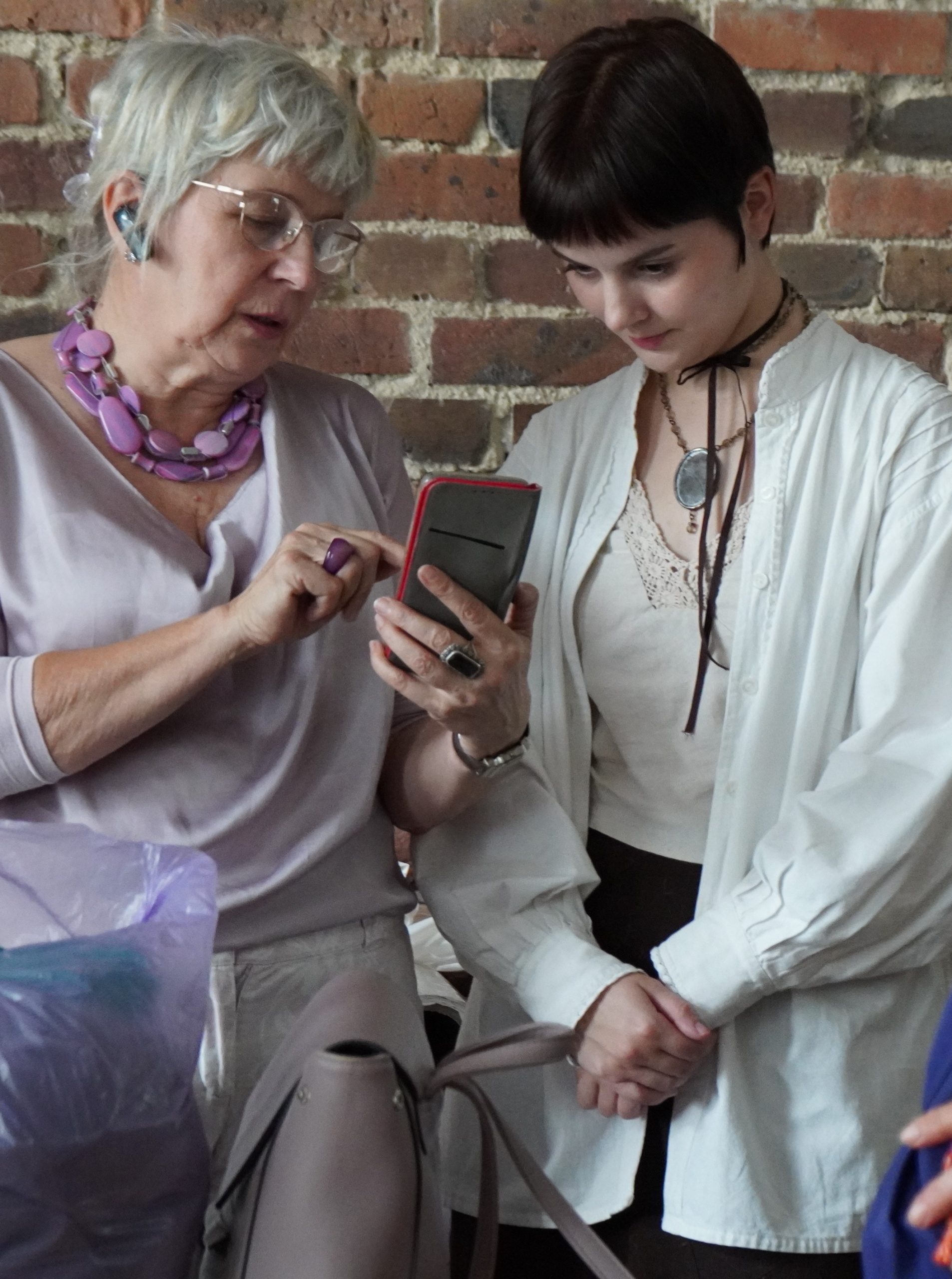 Po lewej stronie stoi starsza kobieta, która trzyma w ręku telefon - coś w nim pokazuje i ma otwarte usta. Po prawej stronie zdjęcia stoi młoda kobieta, która przygląda się w ekran telefonu trzymanego przez starszą kobietę.