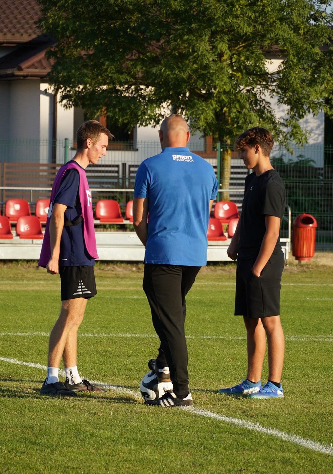 Trzy osoby stojące na boisku sportowym, w tle widać trybuny.