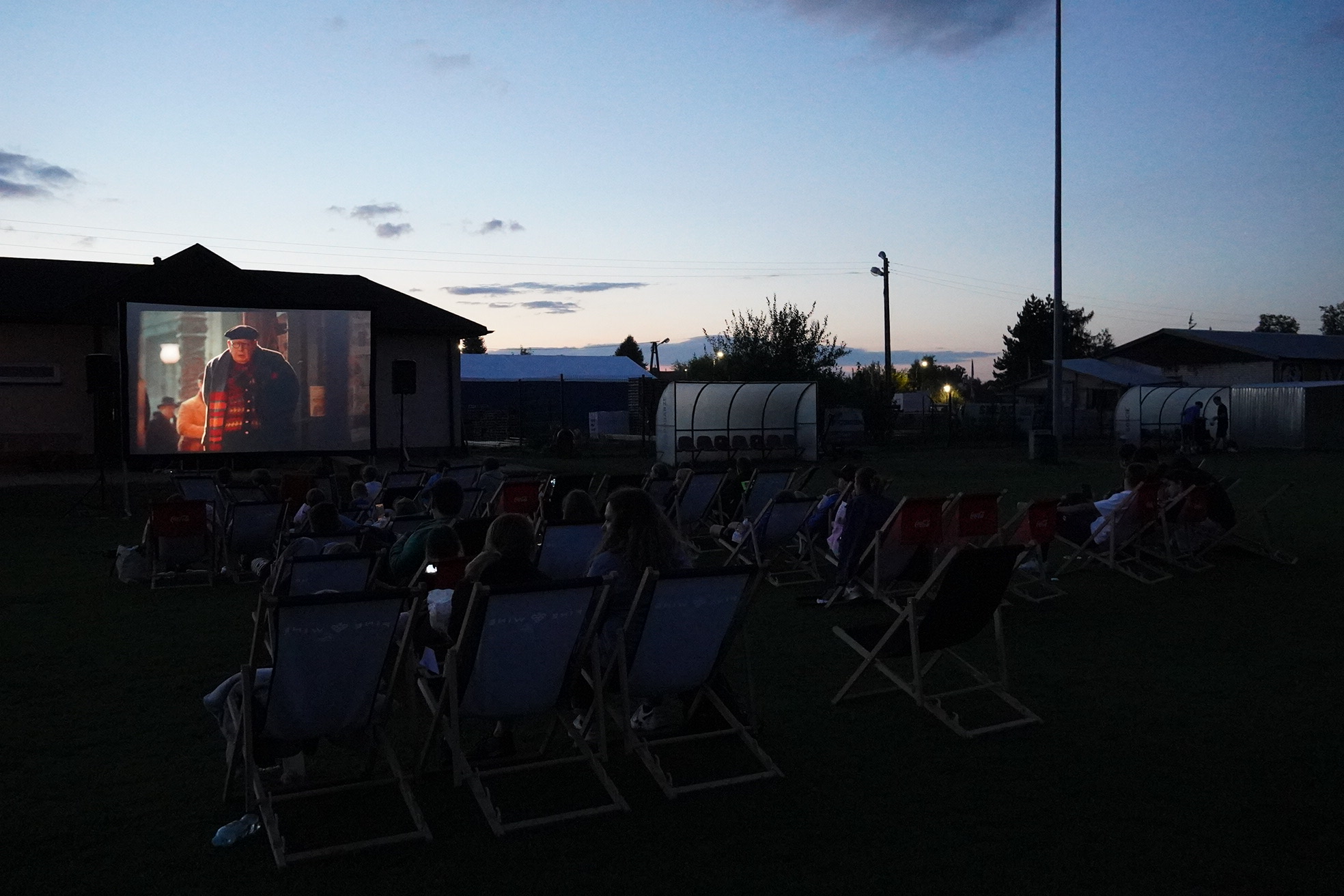 Ujęcie z kina plenerowego - widać ekran, na którym wyświetlany jest film, a także leżaki, na których siedzą widzowie. Zdjęcie jest wykonane o zmroku - jest ciemno, niebo tylko trochę rozjaśnione promieniami niewidocznego już słońca.