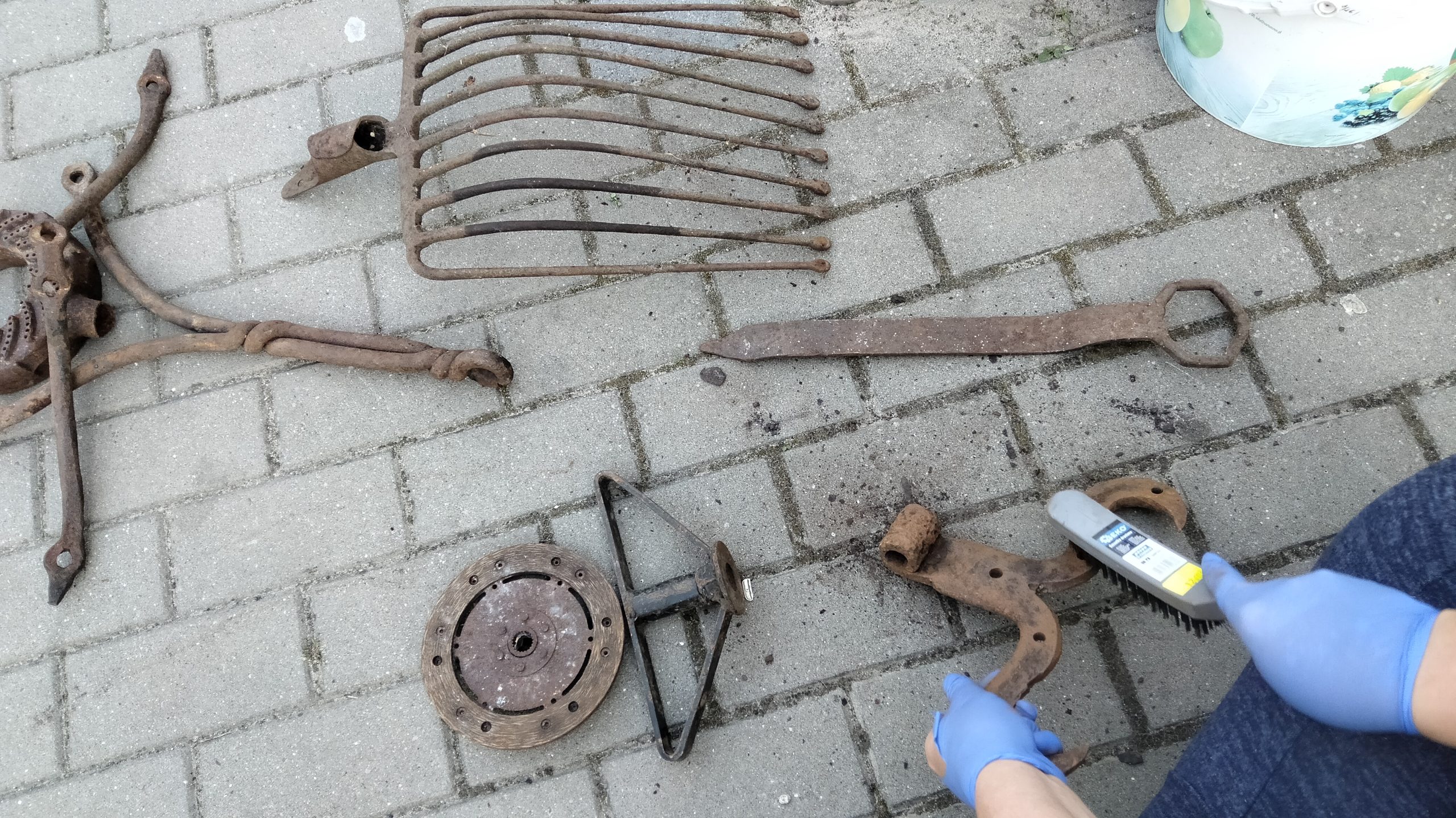 Na chodniku są porozkładane różne metalowe przedmioty, m.in. zawias i grabie.