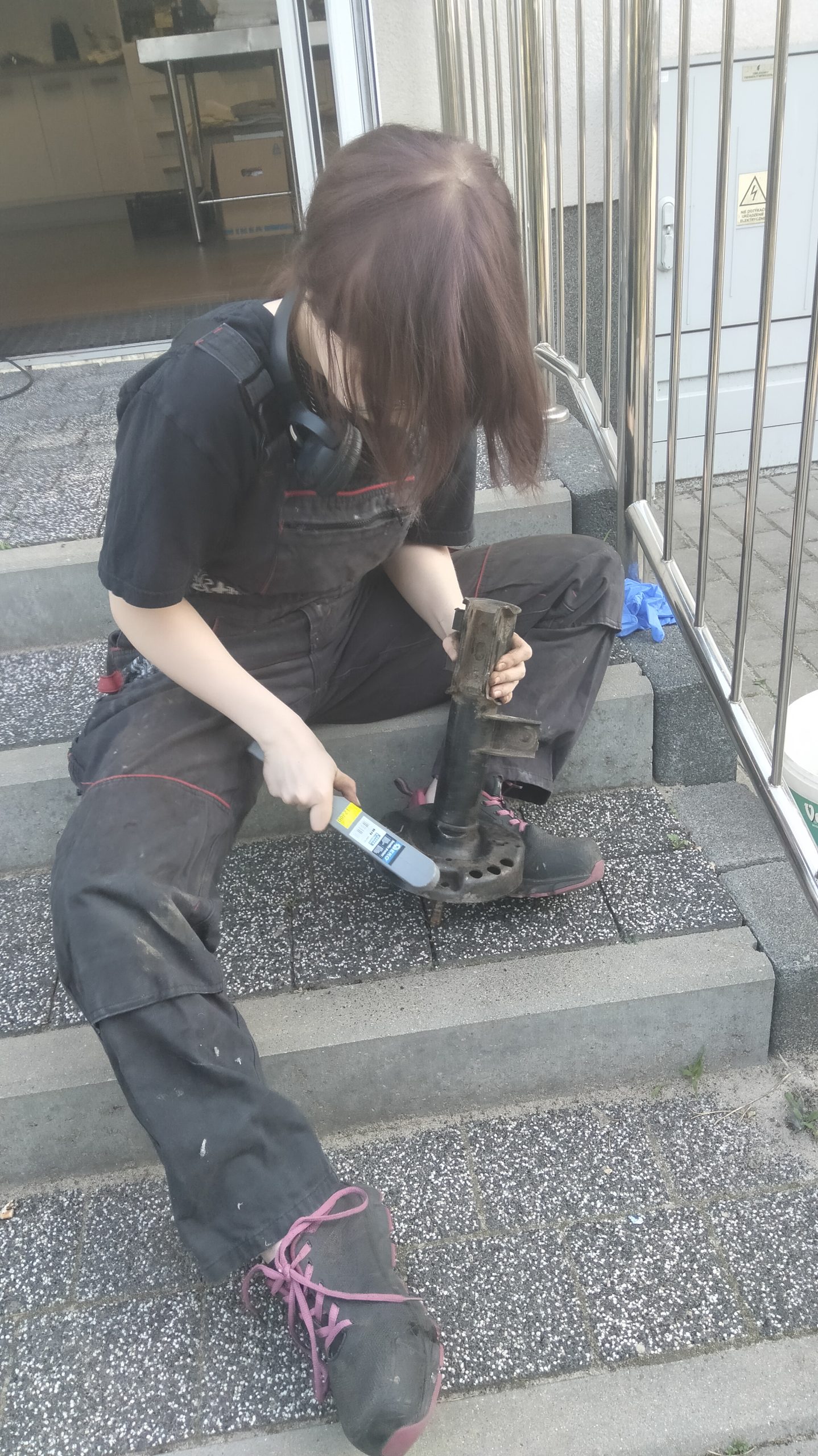 Na schodach siedzi ubrana na czarno dziewczyna, która czyści szczotką duży metalowy przedmiot.