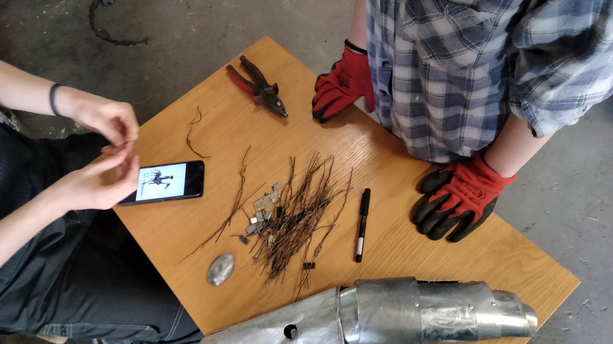W centralnej części zdjęcia jest stół, na którym leżą metalowe kawałki drutu, obcęgi, marker i inne drobne przedmioty oraz włączony telefon komórkowy. Przy stole stoi osoba w koszuli w kratę i rękawiczkach typu 