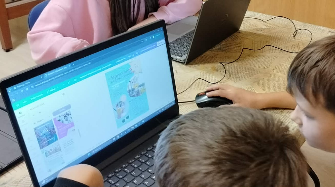 Największą część zdjęcia zajmuje włączony laptop. Na monitorze wyświetla się obraz. Przy laptopie siedzi dwóch chłopców. W tle znajduje się drugi laptop oraz pracująca przy nim dziewczynka.