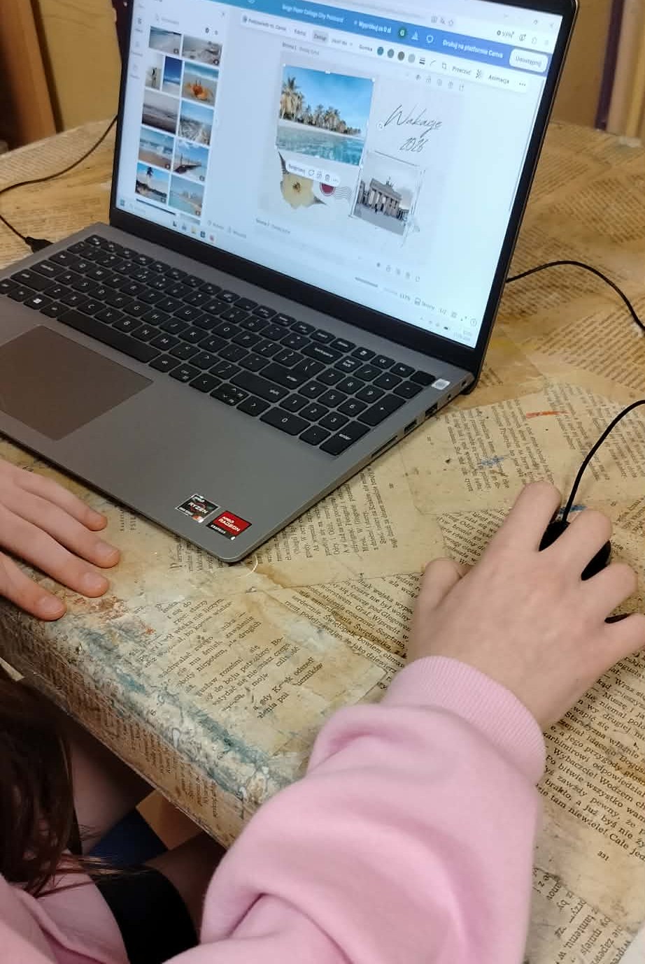 Na zdjęciu widać ręce, jedna ręka trzyma mysz komputerową, druga ręka ułożona przy laptopie na stole. Laptop jest włączony, wyświetla się na nim obraz tworzony w programie komputerowym.