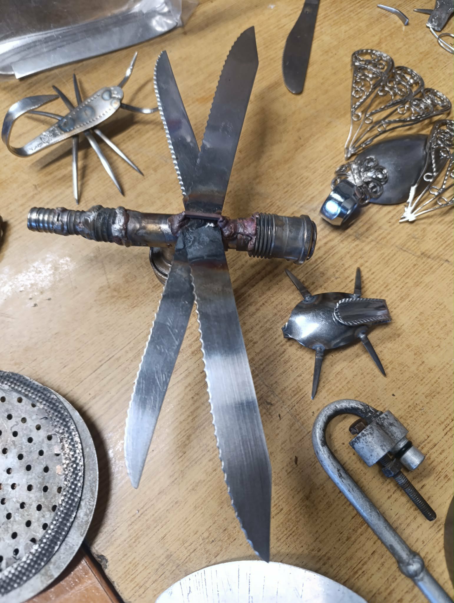 Gotowe prace, wykonane z metalowych fragmentów różnych przedmiotów. Wszystkie przypominają różnego rodzaju owady - m.in. ważka i chrabąszcze. Wszystkie leżą na brązowym blacie.