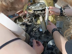 Na zdjęciu po lewej stronie widoczny jest fragment ludzkiej sylwetki, po prawej stronie dwie dłonie trzymające metalowy przedmiot, w centralnej części zdjęcia są różne metalowe drobiazgi.