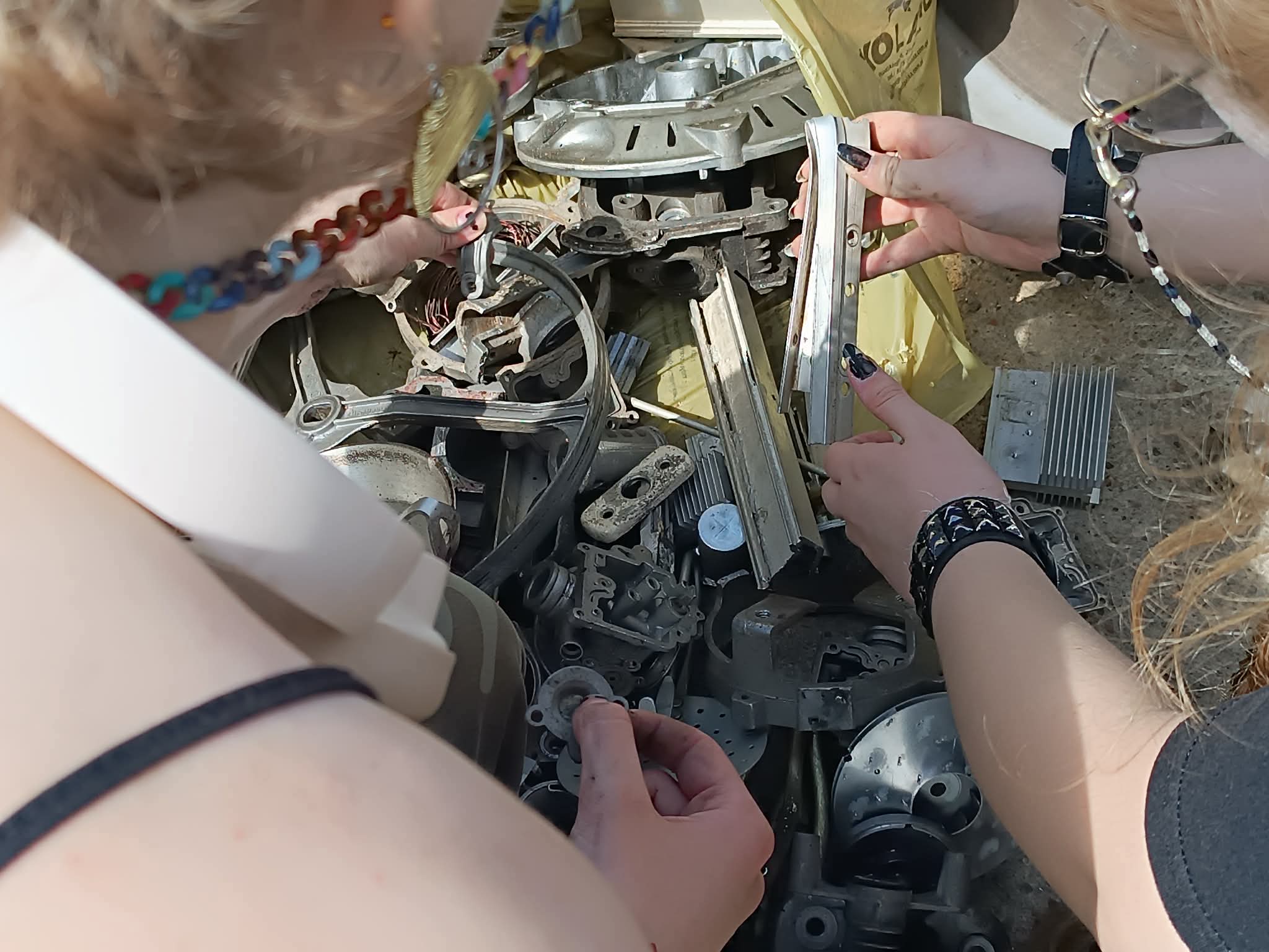 Na zdjęciu po lewej stronie widoczny jest fragment ludzkiej sylwetki, po prawej stronie dwie dłonie trzymające metalowy przedmiot, w centralnej części zdjęcia są różne metalowe drobiazgi.