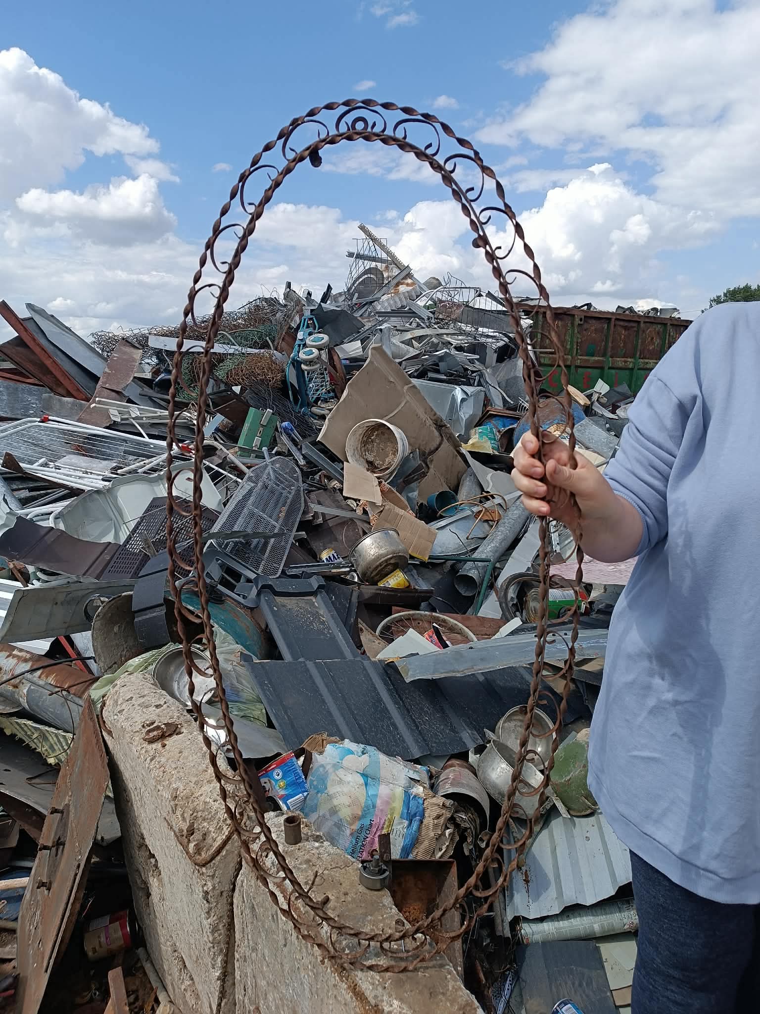 W centralnej części zdjęcia znajduje się metalowa, zardzewiała rama w owalnym kształcie. Po prawej stronie zdjęcia widoczny jest fragment ludzkiej sylwetki - osoby, która tę ramę trzyma. W tle widać bardzo dużo metalowych przedmiotów.