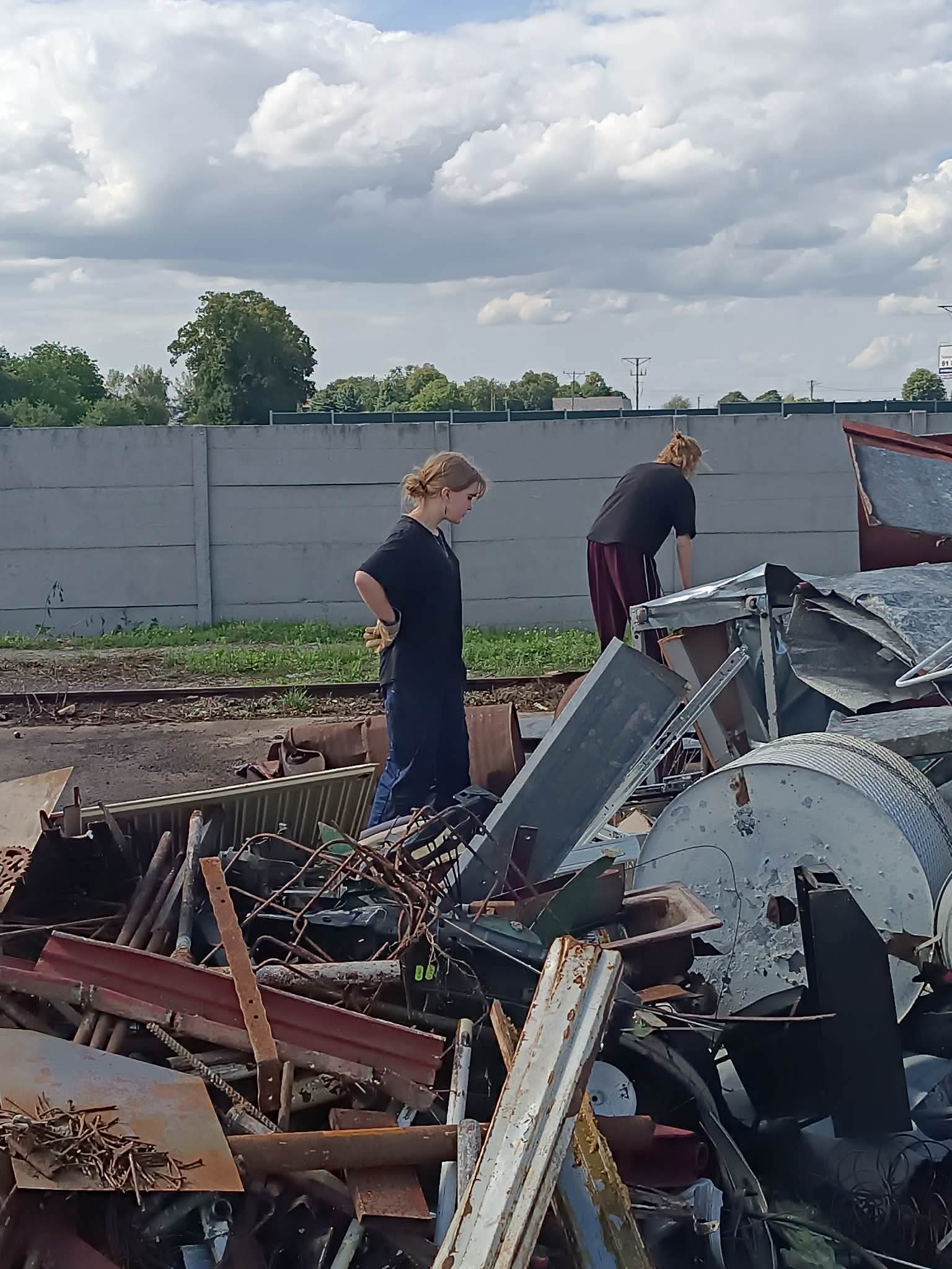 W centralnej części zdjęcia widać dziewczynę stojącą bokiem - patrzy na duże metalowe przedmioty i jest podparta pod boki. Z prawej strony zdjęcia widać odwróconą tyłem, pochyloną osobę, a także dużo metalowych przedmiotów. W tle jest betonowe ogrodzenie, zza którego widać wierzchołki drzew.