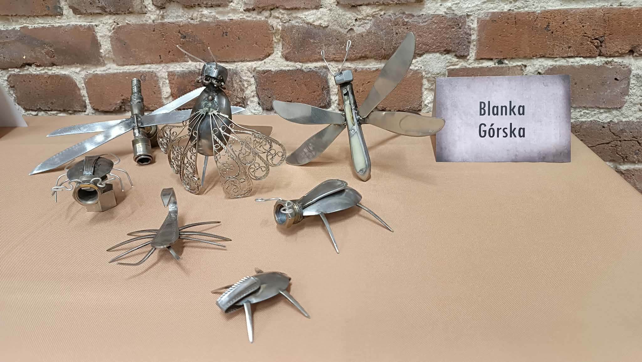 Zdjęcie przedstawia figurki przypominające owady, m.in. żuki i ważki, wykonane ze sztućców i innych metalowych elementów. Obok figurek znajduje się karteczka z napisem