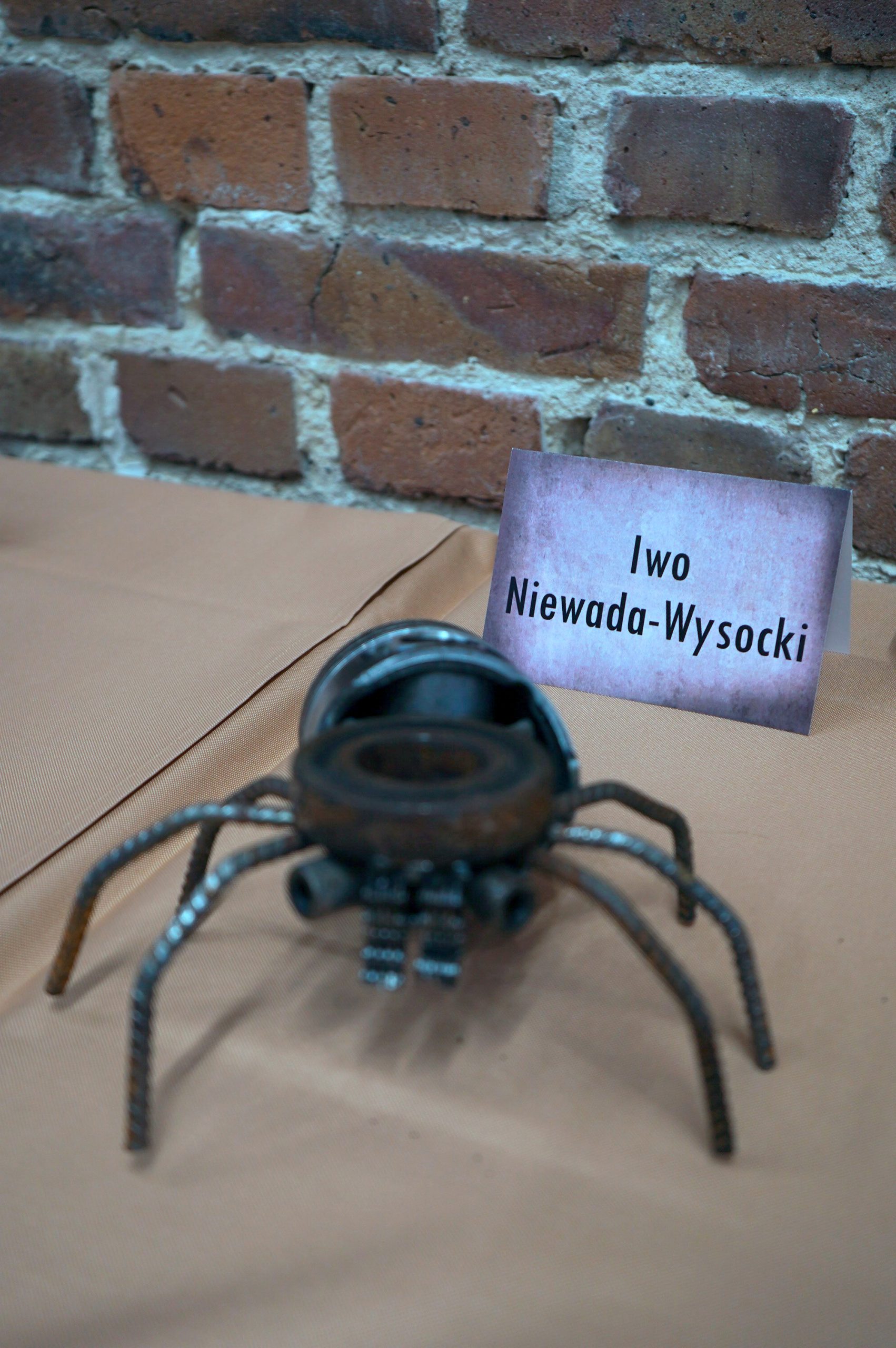 Zdjęcie przedstawia figurkę przypominającą dużego pająka. Figurka została wykonana z metalowych elementów, m.in. drutów, śrub i nakrętek. Obok figurki ustawiono karteczkę z napisem