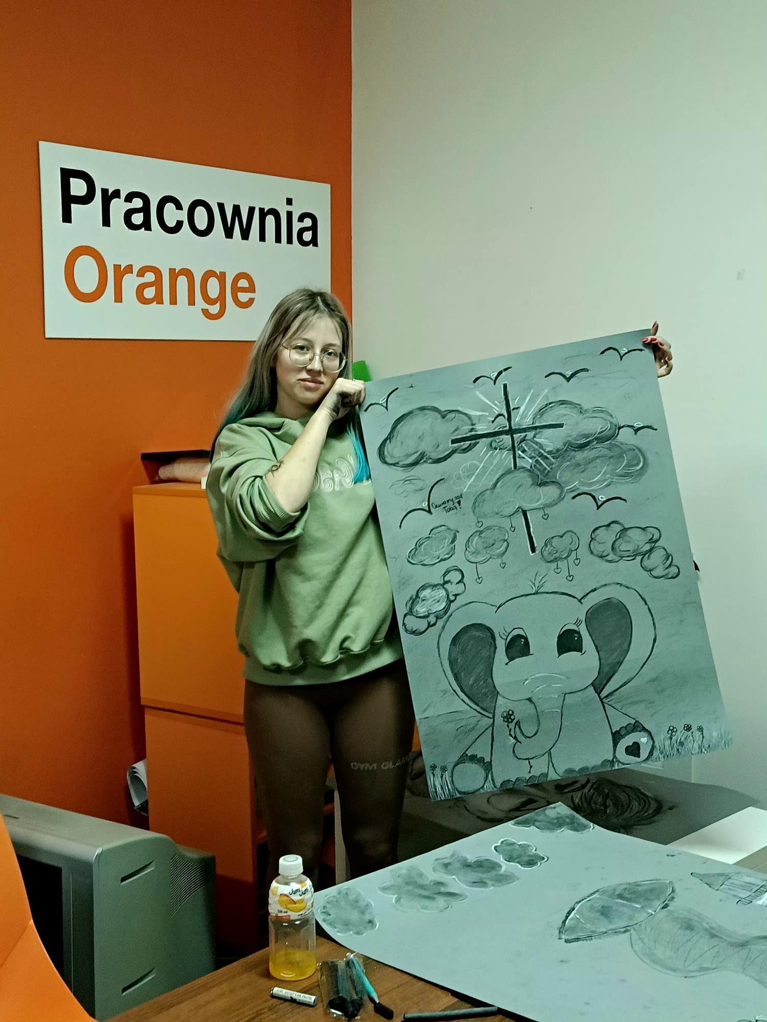 Koordynatorka projektu młodzieżowego, Wiktoria Grzecznowska, prezentuje swoją pracę narysowaną węglem. Obok niej leżą inne dzieła uczestników projektu "Malowanie emocjami".