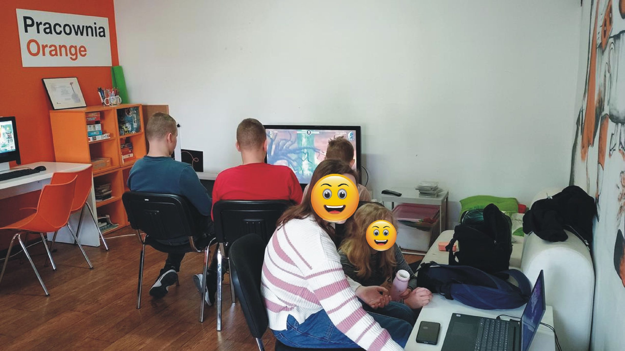 zdjęcie z wnętrza pracowni orange, młodzież gra w grę na playstation, obok dwie osoby oglądają cos na laptopie