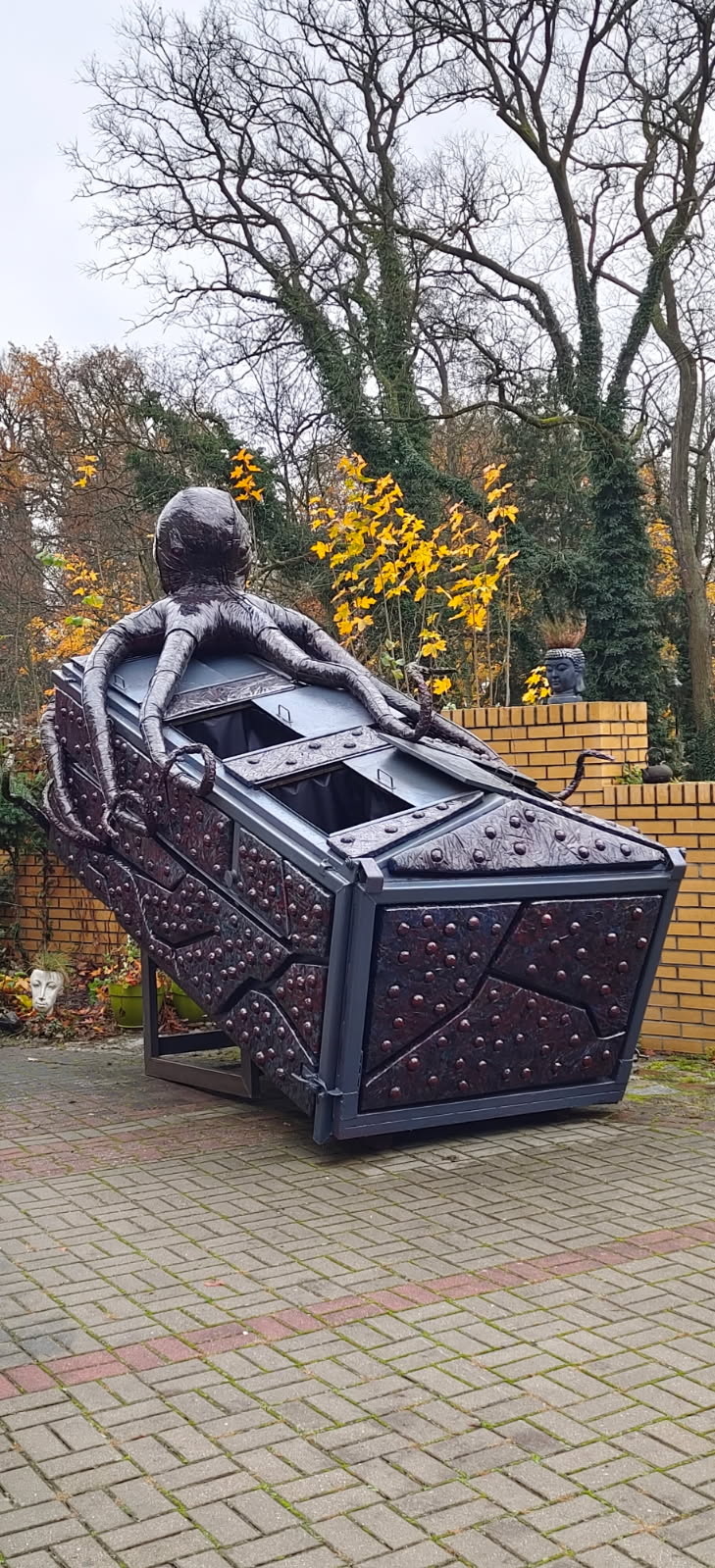 Na zdjęciu widać stary kontener na śmieci, którego obejmuje swoimi mackami wielka ośmiornica. Kontener przyozdobiony jest materiałem identycznym do tego, z którego zrobiona jest ośmiornica. Wszystkie materiały były śmieciem, który zamiast trafić na wysypisko, ozdabia teraz przestrzeń publiczną.