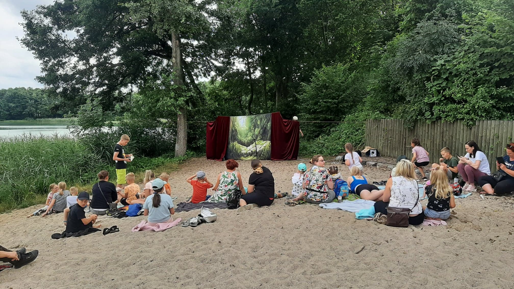 Zdjęcie uczestników i uczestniczek widowiska. Wszyscy siedzą na piasku przed kurtyną zawieszoną na sznurku.