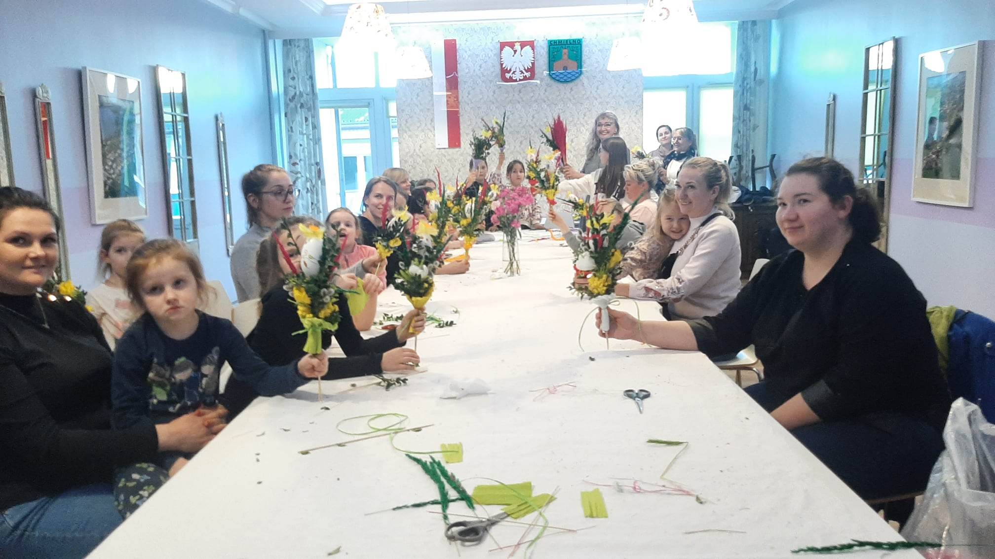 Grupa osób w różnym wieku siedząca przy stole, wyplatająca Palmy Wielkanocne