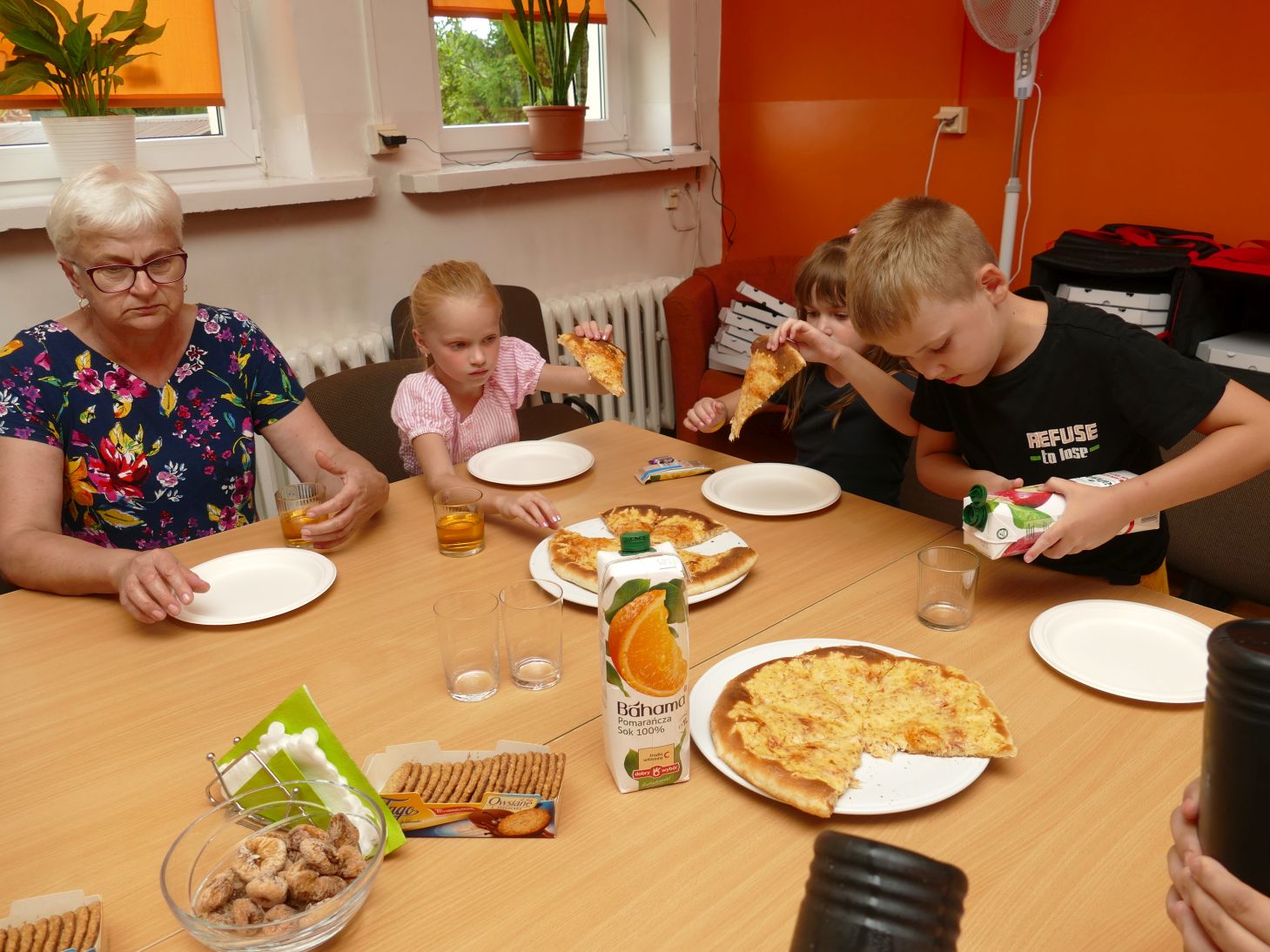 dzieci i starsza kobieta siedzą przy stole i jedzą wspólnie pizze