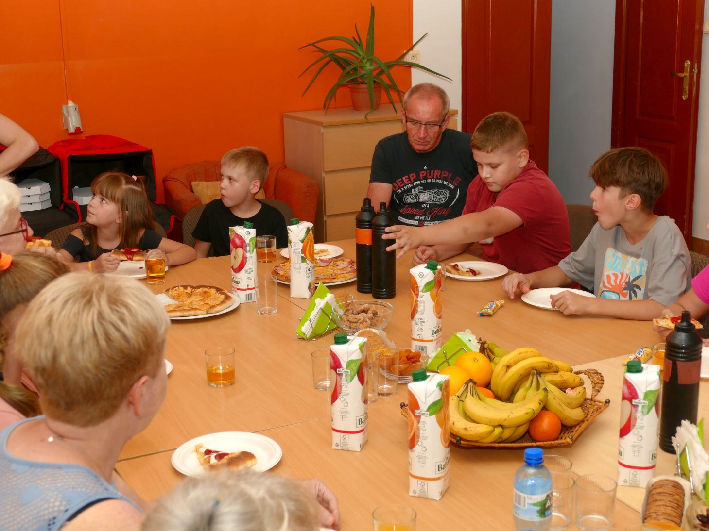na zdjęciu dzieci i starszy mężczyzna siedzą przy stole, na stole stoją soki oraz owoce