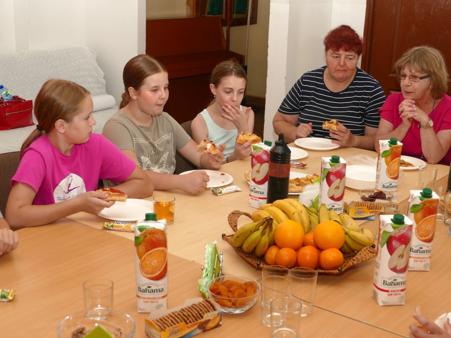 grupa młodych dziewczyn i dwie starsze kobiety siedzą przy stole i jedzą wspólnie