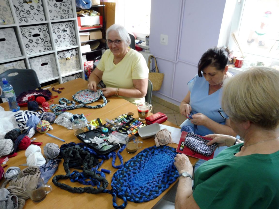 Trzy siedzące kobiety przy stole, wyplatające dywaniki z różnych materiałów