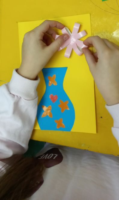 proces tworzenia kartki wiosennej przez dziewczynkę, widzimy jak dokleja do niej jasnoróżowe kwiatki