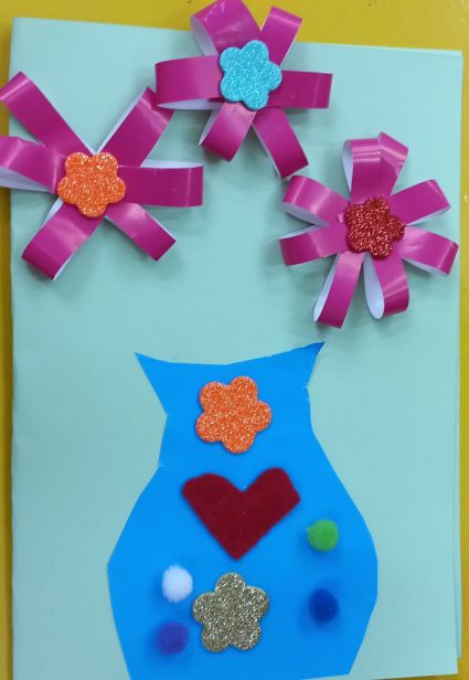 kartka wiosenna z błękitnego papieru, a na niej niebieski wazon, nad nim kwiatki w kolorze fioletowym